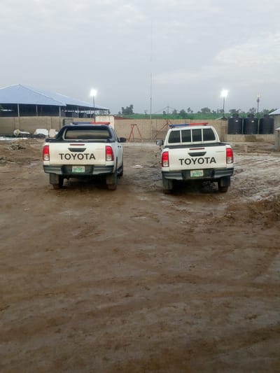 The image shows two white Toyota Hilux pickup trucks parked in an outdoor area that appears to be a construction or industrial site. Both trucks have "TOYOTA" written on the tailgate. The truck on the left has a blue and red light bar on its roof and a green license plate that reads "BWR-704EU". The truck on the right also has a light bar on its roof, though it is less visible, and a green license plate that reads "GWA-991DV". The ground is muddy and uneven. In the background, there is a large building with a blue roof, some large black water tanks, a tall antenna tower, and other construction materials. The sky is overcast and the lighting suggests it is either early morning or late afternoon. The location is identified as Indo, Nigeria.