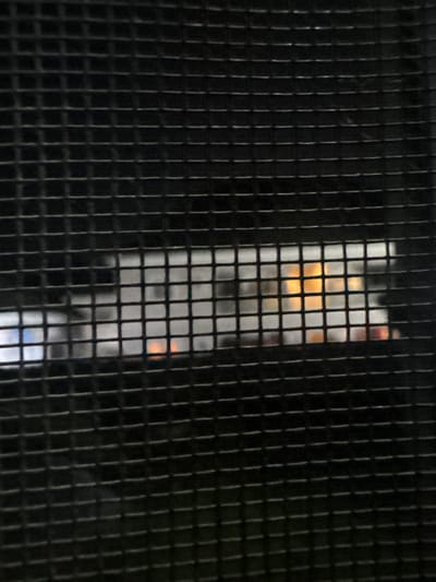 The image displays a close-up view of a black metal grill, obscuring a scene behind it. Through the mesh, a dimly lit interior or outdoor area can be partially discerned. There are faint hints of white structures and a bright orange-yellow light source, suggesting artificial lighting or a flame. The lighting is inconsistent, with some areas appearing brighter than others. The overall impression is that of looking through a barrier at a distance. The location in Angwan Sayawa, Nigeria, is not visually discernible from the provided image.