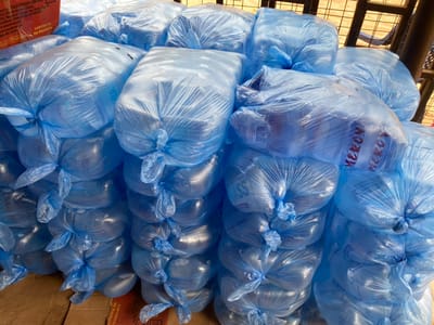 The scene, located in Okura, Nigeria, displays a large quantity of packaged drinking water. Numerous bundles of water sachets are stacked across the frame, forming multiple rows and layers. Each bundle consists of individual water sachets encased within a translucent blue plastic bag, tightly tied at the top. The clear water sachets are visible through the blue plastic. Some bundles reveal text on the contained sachets, including "MERCY" and "CLASS". The stacked bundles rest upon cardboard boxes or sheets, partially visible at the base of the image. A red box with legible text, identifying "TALE FOODS LIMITED" and "Oyo State", along with "CONTENTS: 20 Packets", is partially visible on the far left. No individuals are present. The background shows a partial view of an indistinct structure, possibly a metal frame, suggesting an indoor or covered storage environment.