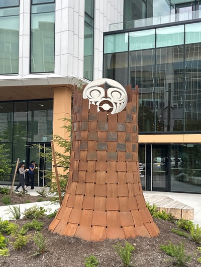 This image depicts an outdoor scene in what appears to be an urban environment in Electoral Area A, Canada. The main feature is a large, woven sculpture made of reddish-brown rectangular tiles, positioned in front of a modern building with a glass facade. The sculpture features a white circular element with a stylized design on top. Two individuals are visible in the scene, standing near the base of the sculpture and the building’s glass walls. They appear to be looking at their phones. The ground around the sculpture is covered in mulch and some green plants. The environment suggests it is a public space, likely near an office or commercial building.