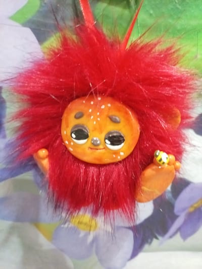 This is a close-up, eye-level, vertical, indoor shot of a handmade fuzzy creature. The creature has a round, orange face with a small smile, large, cartoonish black and white eyes with white dots, and a tiny nose. The face is adorned with small white dots. A fluffy, bright red mane surrounds the face, giving it a lion-like appearance. The creature has two orange arms and two orange feet, with one foot holding a small bee-shaped charm. The background is a blurry fabric with a floral pattern featuring purple and green hues. The lighting suggests it's daytime, and the setting appears to be a home environment in Bologoe, Russia. The overall emotion conveyed is whimsical and playful. There is no visible text in the image.