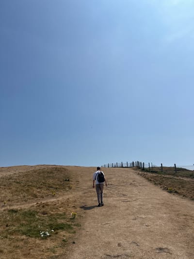 A person walks up a dirt path on a dry, grassy hill under a clear blue sky. The path is wide and leads towards a wooden fence that runs along the top of the hill. The person is wearing a white t-shirt, beige pants, and a black backpack. The grass on the hill is mostly brown and dry, with patches of green and some small yellow and white wildflowers. In the distance, a body of water is visible to the right of the path. The image appears to have been taken on a sunny day. This scene is consistent with the landscape found at Seaford Head, Seaford, United Kingdom, known for its chalk cliffs and coastal paths.