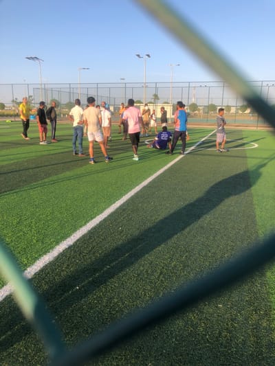 The image displays an outdoor artificial turf soccer field located in النعيلة, Saudi Arabia. Approximately fifteen to twenty adult males are present on the bright green turf, viewed partially through a foreground wire mesh fence and a prominent, blurred diagonal green bar. The field features distinct white markings, including a solid white line running diagonally from the lower left towards the mid-right, and a segment of a circular arc. Most individuals are standing in small clusters or individually across the mid-ground, dressed in various casual attire including t-shirts, shorts, and long pants. One male, wearing a blue jersey with the number '22' on his back, is seated on the turf with his legs extended. The individuals appear to be engaged in casual social interaction, or are taking a break from or preparing for a sports activity. Long, dark shadows are cast across the field from the individuals and other structures, indicating a low sun angle, consistent with either early morning or late afternoon. In the background, beyond a perimeter fence, several tall light poles equipped with solar panels and floodlights are visible, along with sparse vegetation and distant low-rise buildings under a clear, pale blue sky.
