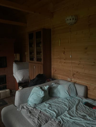 The image shows the interior of a rustic wooden cabin room. A large, light gray sofa is made up as a bed and covered with teal and gray bedding. Two large teal pillows are on the bed. A black backpack is on the floor behind the sofa. To the left, a red brick fireplace is visible, with a black mantelpiece and a dark hearth. Above the fireplace, a dark framed object is mounted on the wall. A tall, wooden cabinet with glass doors and wooden shelves is situated behind the sofa on the left. A white cushioned chair is leaning against the cabinet. The walls and ceiling are paneled with light-colored wood. A white, globe-shaped light fixture with dark circular accents hangs from the ceiling on the right side of the room. A white cord runs down the wall from the light fixture to an on/off switch and then to electrical outlets. A thermometer is mounted on the wooden wall. The floor is wooden and partially covered by a dark rug. A video game controller with red and blue accents lies on the bedding on the right side of the bed. The overall impression is of a lived-in, cozy space. The location is described as Красное, Russia, suggesting this is a residential interior.