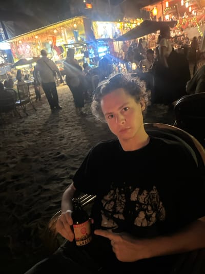The image depicts a nighttime scene at a beachside establishment in Kolamb (Kollam), India. The foreground features a light-skinned male, estimated to be between 20 and 30 years of age, with short, dark, curly hair, seated in a wicker-style chair. He is wearing a black t-shirt with a detailed, light-colored graphic print on the chest. His left hand firmly grips a dark glass bottle with a partially visible label identifying it as 'LAGER.' His left index finger points directly at this bottle.

The background shows a densely populated outdoor area with a sandy ground texture. Multiple temporary structures, resembling open-air restaurants or shacks, are brightly illuminated by string lights, red lanterns, and blue neon lighting. Numerous individuals, casually dressed, are visible throughout the background, engaged in walking, sitting at tables, and social interaction. Additional wicker-style chairs and tables are present, some beneath awnings or umbrellas. Artificial light sources are the primary illumination.