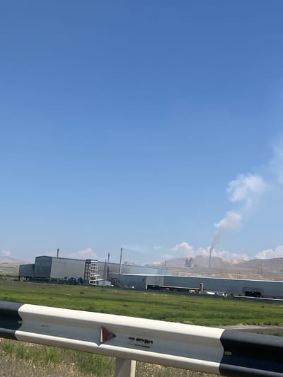 The image shows a landscape scene with an industrial facility. The sky is clear and blue with some white clouds. In the distance, a factory complex with several buildings and smokestacks is visible. One smokestack is emitting a plume of dark smoke, suggesting industrial activity. The foreground is dominated by a white and black barrier, possibly a guardrail, with some rust and a small red and white reflector. To the left of the barrier, a green grassy field stretches towards the industrial buildings. The overall impression is of a bright, sunny day in an industrial area. There is some faint, illegible text on the barrier. The image appears to be taken from a roadside perspective, looking towards the industrial site and the sky. The scene is likely in Surenavan, Armenia.