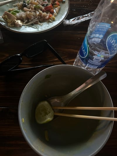 The scene, located in Ban Paem Bok, Thailand, displays the aftermath of a meal on a dark brown wooden table. In the foreground, a light grey ceramic bowl contains a metal spoon, two wooden chopsticks, and a squeezed lime wedge, with minimal yellowish residue at the bottom. To the left of the bowl, a pair of black sunglasses with dark lenses lies open on the table. A clear plastic bottle of "CRYSTAL" brand water, featuring Thai script, is positioned diagonally to the right of the sunglasses and behind the bowl. In the background, a light green ceramic plate holds remnants of a dish primarily composed of rice, chicken pieces, and various chopped vegetables, including green segments and red tomato fragments. A metal fork with a golden-colored handle rests within the food on the plate. No individuals are present.