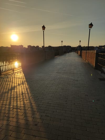 The image depicts a sunset over the city of Badajoz, Spain, looking east across the Puente de Palmas. The sun is low on the horizon, casting long shadows from the bridge's railing onto the cobblestone walkway. The Puente de Palmas is a historic bridge spanning the Guadiana River, characterized by its brick parapets and numerous lampposts. In the distance, the cityscape of Badajoz is visible, with buildings silhouetted against the warm, golden sky. Several individuals are walking on the bridge, enjoying the evening light. The scene is serene and picturesque, capturing the beauty of the city at dusk.