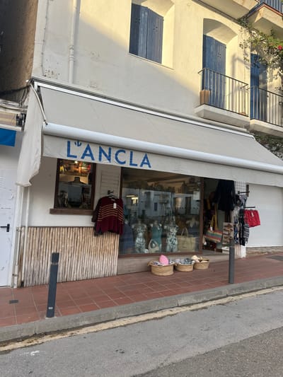 The image depicts the storefront of a shop named "L'ANCLA" located in Calella De Palafrugell, Spain. The building features a white stucco facade with several windows, including two on the upper level with closed blue wooden louvered shutters and a third on the right with a small black iron balcony. A second-floor balcony with a similar railing is visible further right, partially obscured by green foliage with red flowers.
The ground floor storefront is covered by a retractable beige awning. Below the awning, the shop's name, "L'ANCLA," is prominently displayed in blue capital letters. The shop window displays several items, including three headless mannequins wearing patterned swimwear and various bags and accessories. Inside the shop, reflections from the opposite side of the street are visible in the glass.
To the left of the main display window, a smaller window is present, with a male torso mannequin visible inside. Below this window, the lower portion of the wall is clad with vertical light-colored wooden or bamboo slats. A dark-colored garment with horizontal red and beige stripes hangs on a hook outside the shop, to the left of the main display.
To the right of the main shop window, a black metal grid rack holds various clothing items, including patterned tops and red shorts. Directly in front of the shop window, on the tiled terracotta sidewalk, three circular woven baskets contain merchandise such as striped cushions and hats.
The sidewalk is paved with terracotta-colored tiles, leading to a grey concrete curb. A black and grey bollard is fixed into the sidewalk near the left side of the frame. Beyond the curb, an asphalt road with visible wear and light colored markings is present. The scene is illuminated by strong sunlight from the right, casting shadows on the building and highlighting textures. No individuals are visible in the frame.