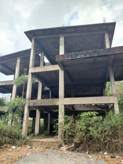 The image depicts an unfinished concrete building structure, likely abandoned, in Marna, India. The structure consists of multiple stories of reinforced concrete pillars and beams, without walls or windows. Some reinforcing bars protrude from the concrete edges. Vegetation has grown around and through the structure, obscuring parts of the lower levels. Debris and trash are scattered on the ground around the base of the building. The sky is overcast.