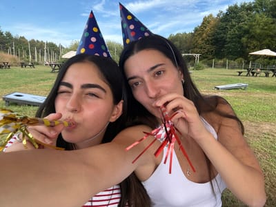 The image displays two young adult females outdoors in Accord, United States, engaged in a celebratory activity. Both individuals wear dark blue party hats with multicolored polka dots. The female on the left, wearing a red and white striped top, winks with her left eye and blows into a gold and yellow party blower, holding it with her left hand extended, indicating a selfie. The female on the right, wearing a white tank top with a silver necklace, blows into a red and white party blower. The background features a grassy area under a blue sky with scattered clouds. Visible objects include multiple wooden picnic tables with benches, two cornhole boards on the grass, two large beige patio umbrellas, wooden posts, a wooden arbor, and distant trees with some autumn foliage.