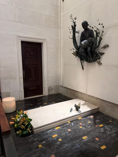 The image depicts an interior corner of the Basilica of Our Lady of the Rosary, located within the Sanctuary of Fátima in Fátima, Portugal.
The scene features the tomb of Saint Francisco Marto. This rectangular, light-colored marble slab, inscribed with text (partially showing "FRANCISCO"), rests on a dark, polished stone floor with scattered, irregular square inlays of yellow-orange and reddish-brown hues. On the tomb slab are two small, dark-colored metallic bird figurines; one larger with spread wings, the other smaller and stationary. At the left end of the tomb, a floral arrangement consisting of orange lilies and green foliage rests on the floor. Adjacent to the flowers, a large, cylindrical, translucent object, possibly a stone lantern or candle holder, emits a soft, warm light against the light-colored wall.
To the left, a dark brown wooden door with recessed, carved geometric panels and a vertical metal handle is set within a plain, light-colored stone doorway. The walls throughout the scene are constructed from large, uniformly cut, light-colored stone blocks. Mounted on the right-hand wall, above the tomb, is a bronze sculpture depicting a young shepherd boy seated amidst stylized tree branches and leaves, cradling a lamb. The sculpture includes a shepherd's staff and smaller bird and leaf elements. No individuals are present in the immediate scene.