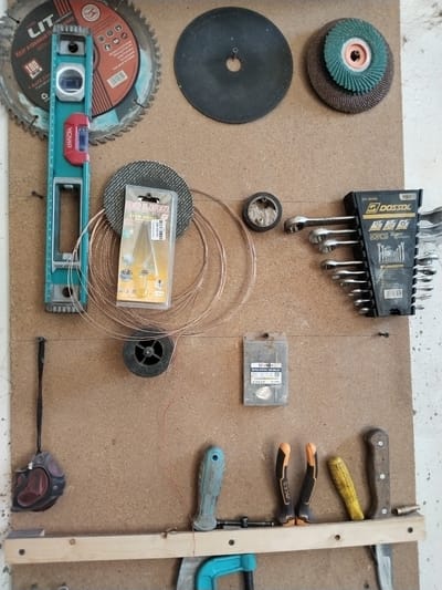 This image shows a wall-mounted tool organizer in Chalysh, Uzbekistan. The organizer is made of corkboard and displays a variety of tools.
In the top left corner, a circular saw blade with the brand "LIT" is visible. To its right is a black abrasive disc. Further to the right, a green flap disc is mounted on a brown abrasive disc.
Below the saw blade is a turquoise level with red accents. To its right, a small package of "BOLIAN" brand step drill bits is attached to the board. A bundle of copper wire is coiled below this package.
Further down, a small black spool with red wire wound around it is mounted. To the right of this spool is a black organizer holding a set of "DOSSOL" wrenches. Below the step drill bits, a small plastic package labeled "VIBRO" containing what appears to be drill bits is also present.
On the bottom shelf, from left to right, are a retractable tape measure, a blue handled tool, two black and orange handled tools, a yellow handled tool, and a wooden handled tool. Other smaller tools and fasteners are also visible. The background appears to be a concrete wall. The scene depicts a tidy storage system for tools.