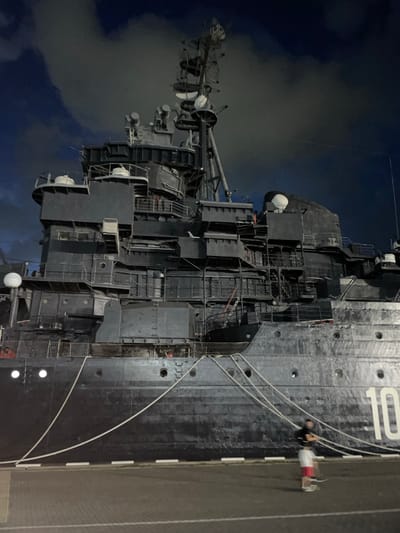 The image shows a large, dark-colored ship docked at night in Verkhnyaya Stavropol'ka, Russia. The ship has a complex superstructure with multiple levels, antennas, and equipment. White mooring ropes are visible, securing the ship to the dock. A single person, dressed in red and white shorts and a dark t-shirt, is visible in the foreground, appearing to be walking or standing near the ropes. The sky above is dark and cloudy. The hull of the ship displays the number "10" in large white numerals.