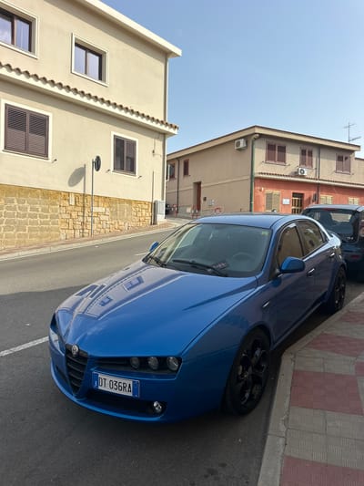 The image shows a blue Alfa Romeo sedan parked on the side of a street in Crabonaxa, Villasimius, Italy. The car has a license plate that reads "DT 036RA." The street is paved with asphalt and has a sidewalk made of red and beige tiles. In the background, there are two buildings. The building on the left is a two-story structure with a beige facade and brown-shuttered windows. The building on the right is also two stories, with an orange-colored facade and brown-shuttered windows. The sky is clear and blue, indicating a sunny day. The scene appears to be a residential area. There are no people visible in the image.