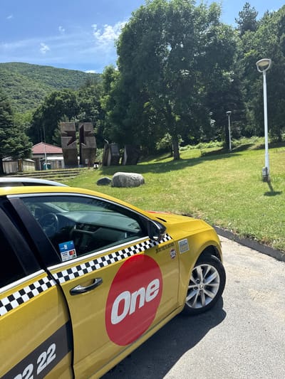 The image displays an outdoor scene featuring a yellow taxi cab parked on an asphalt surface. The taxi, possibly a Skoda given the wheel design, has a prominent red circle with "One" in white letters on its side and a black and white checkered stripe running along its length. Visible text includes payment card logos such as "VISA," "Mastercard," "Postbank," and a contactless symbol on a sticker near the driver's window. The number "22" is partially visible on the rear side of the car.
To the left of the car, a lush green grassy area is bordered by several large, leafy trees. Two modern streetlights stand within the grass. Further back, an abstract sculpture made of dark, block-like structures and a large grey rock are positioned on the lawn. A residential building with a red-tiled roof is partially visible behind these elements. In the distant background, a sprawling forested mountain range rises under a clear blue sky dotted with a few white clouds.
The strong sunlight and distinct shadows suggest it is midday or early afternoon on a clear, sunny day. No people or activity are visible, creating a serene atmosphere. The location cues, including the specific taxi branding and the natural landscape, place the scene in Vetren Dol, Bulgaria.