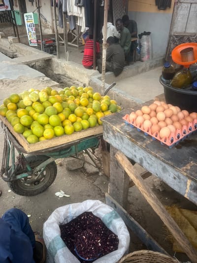 An outdoor market scene shows a variety of goods for sale. A cart overflowing with green citrus fruits sits beside a tray of eggs. A large white bag filled with dark red flower petals is positioned nearby. In the background, several individuals are seated, and various objects suggest a bustling market atmosphere. The photo captures the diversity of goods available in the market setting.