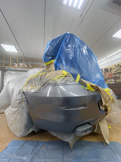 The image depicts the rear bumper of a car, which has been covered with blue plastic sheeting and masking tape for protection. The bumper itself is a dark grey color and appears to be freshly painted or prepped for painting. A portion of a white plastic wrap is visible on the left side, covering more of the vehicle. In the background, there are shelves with boxes, tools mounted on a wall, and fluorescent lighting fixtures on the ceiling. The floor appears to be a cork or linoleum material, with a blue plastic sheet placed in the foreground. There are no people visible in the image. The location is identified as Veteran-2, Russia.