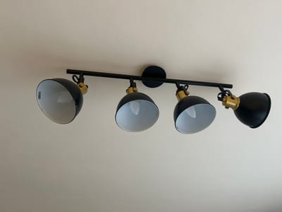 This media file displays a four-light track light fixture mounted on a textured, off-white ceiling. The fixture itself is black with brass accents, featuring adjustable spotlights. Each spotlight has a black exterior and a white interior, with a light bulb visible inside. The fixture is centrally located in the frame, with a significant portion of the ceiling visible above and below it. There is no indication of the time of day, weather, or specific location within Limerick, Ireland, beyond the fact that it is an indoor setting. No people are present in the image. The overall scene is static and focuses solely on the lighting fixture and the ceiling. There is some faint text visible on the inside of the leftmost spotlight, but it is too small and blurry to be legible.