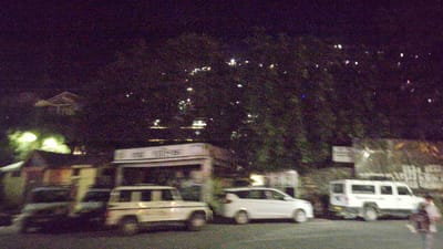 The image depicts a nighttime street scene in Kullu, India, characterized by a dark sky dotted with numerous lights from buildings and potentially distant stars or atmospheric phenomena. In the foreground, several vehicles are parked or moving. A white SUV is on the right side of the road. To its left, a white, older model utility vehicle is visible. Further left, a white car is parked. A building with a sign is behind the vehicles, suggesting it might be a commercial establishment or a public service building. The overall environment appears to be a built-up area within Kullu, likely a town or city street, with trees and foliage present, illuminated by artificial lights from structures and vehicle headlights. The image is grainy due to low light conditions.