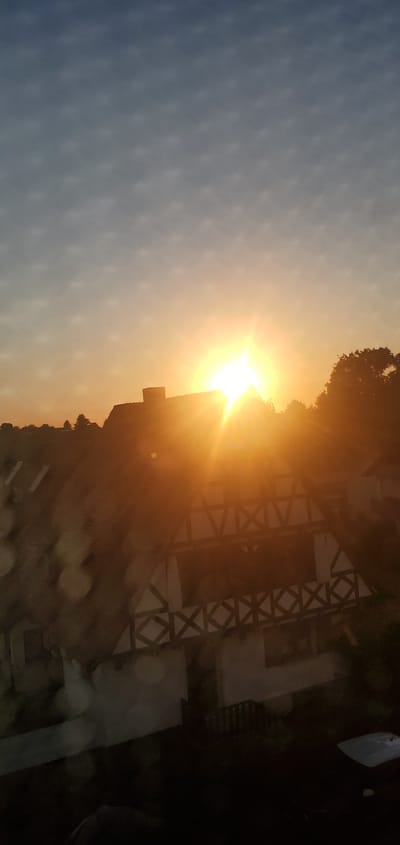 The image captures a sunset over a village in Sasbach, Germany. The sky transitions from a deep blue at the top to a bright orange and yellow as it nears the horizon. A radiant sun, partially obscured by a building's roof, casts strong rays of light across the scene. In the foreground, a traditional half-timbered house with prominent dark beams against white plaster is visible. The house features multiple windows and a balcony or porch with a decorative railing. Trees are silhouetted against the vibrant sky on the right side, and a line of trees also forms the distant landscape. The lower part of the image is darker, with some objects and structures faintly discernible, including what appears to be a car. Water droplets on a surface, possibly a window, create bokeh effects in the lower left portion of the image, suggesting it was taken from indoors. The overall impression is that of a serene, rural evening in Sasbach.