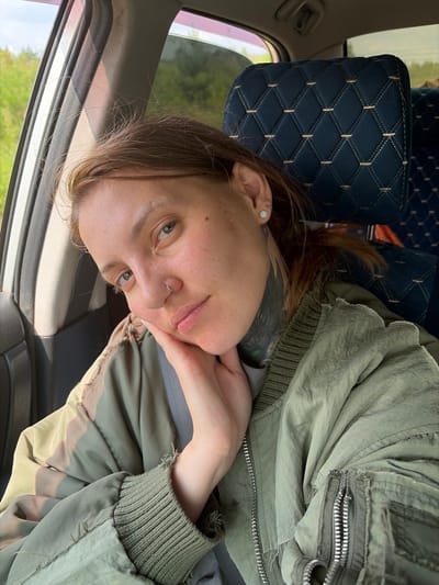 A young Caucasian woman with auburn hair, visible tattoos on her neck, a nose ring, and an earring is looking towards the camera, resting her chin on her hand, from the passenger seat of a vehicle. She is wearing a green bomber jacket. The interior of the car features a dark blue upholstered headrest with a diamond pattern. The background shows a blurred green landscape through the car window, suggesting a rural or natural setting, potentially on the outskirts of Kamennyi Kliuch, Russia. The lighting indicates it is daytime.