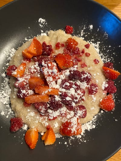 On a black plate, there is a serving of porridge-like dish, topped with diced strawberries and raspberries, dusted with powdered sugar. The dish is served on a wooden surface. The scene does not provide enough details to determine the location in the city of Lommedalen, Norway. There is no information about people, actions, or interactions.