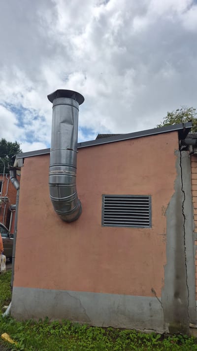 A galvanized metal chimney is attached to the side of a salmon-pink stucco building. The chimney is curved, extending from the roofline and then bending downwards. The building has a gray concrete base and a dark gray roofline. A rectangular ventilation grate is visible on the wall below the chimney. There is a significant crack running down the right side of the building's facade, exposing brickwork beneath the stucco. Overcast, cloudy skies are visible above. Green grass and some weeds grow at the base of the building. A portion of a car and a person are visible to the left of the building. The image appears to be taken outdoors in Jēkabpils, Latvia.