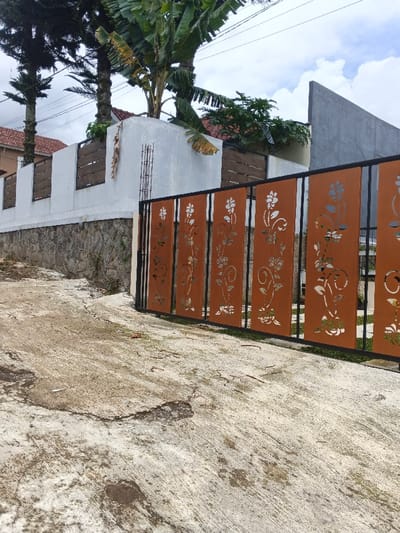 A modern fence borders a property. The fence consists of decorative, rust-colored panels with laser-cut floral designs, set within a black metal frame. Behind the fence, a stone and white wall is visible, topped with a wooden horizontal slat section. Lush trees and other vegetation are seen above and behind the wall. The foreground depicts a cracked, aged concrete driveway leading up to the fence. The sky is overcast.