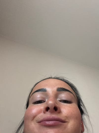The image shows a close-up, low-angle, tilted view of a young woman's face. She has dark hair and appears to be wearing makeup, including defined eyebrows and lip gloss. Her skin has a dewy sheen, suggesting it might be highlighted or moisturized. Her eyes are partially closed, and she has a slight smile on her lips, conveying a relaxed or perhaps playful emotion. The background is a plain, light-colored wall with a subtle texture and a seam running diagonally across it. The lighting appears to be soft and diffused, possibly from an indoor source. The framing and angle of the shot suggest a selfie taken from a unique perspective. There is no visible text in the image. The location context provided is Koltushi, Russia.