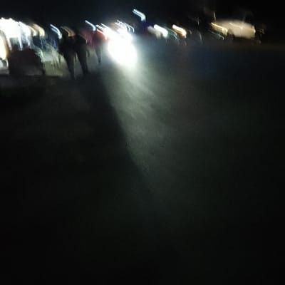 A nighttime street scene displays blurred figures walking on the left. A vehicle's headlights illuminate a path in the center. Light sources flicker in the background, possibly from streetlights or buildings. The overall impression is of motion and limited visibility due to darkness.