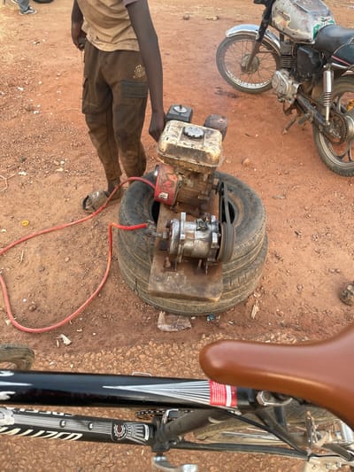 A person is standing in a reddish-brown, gravelly area next to a homemade air compressor. The air compressor is set on top of two stacked tires. The compressor is attached via a belt to a small engine. An orange air hose is connected to the compressor. Next to the compressor is a black bicycle with a brown seat, and in the background is a motorcycle.