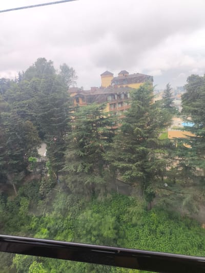 The image shows a view of a building and surrounding trees from what appears to be a cable car in the city of Kutaisi, Georgia. The building is multi-storied with a light yellow facade and a dark brown roof, featuring numerous windows and balconies. It is nestled among dense green coniferous trees, some of which tower over the structure. In the background, more buildings and a glimpse of a blue swimming pool can be seen. The sky is overcast with a pale grey cloud cover. The foreground shows the dark railing of the cable car. The scene is one of a scenic hillside location with a hotel or residential complex situated amidst a verdant, natural setting.