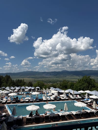 The image depicts a resort with multiple swimming pools and lounge chairs under white umbrellas, set against a backdrop of rolling green hills and a cloudy blue sky. In the foreground, people are seen relaxing on lounge chairs and swimming in the pools. The resort appears to be located in Sapareva Banya, Bulgaria, a town known for its hot springs and natural beauty. The scene conveys a sense of leisure and relaxation, with the clear blue water of the pools and the lush greenery of the surrounding landscape creating a tranquil atmosphere. The mountainous terrain in the distance adds to the picturesque scenery.