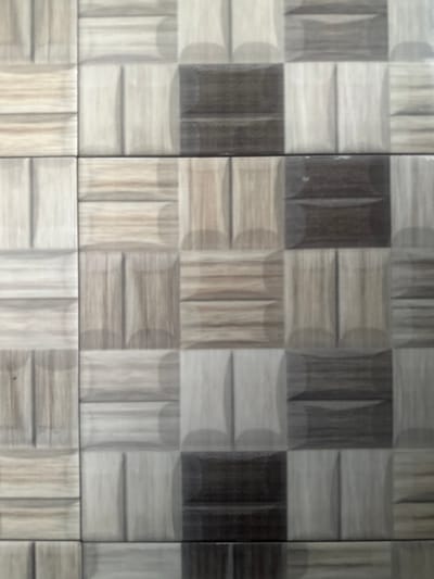 A repeating pattern of square tiles with a dimensional, textured surface. Each tile is divided into four rectangular segments, arranged in a square formation. The segments exhibit variations in shade, ranging from light gray/beige to dark brown. The overall effect is a mosaic-like design with a subtle 3D appearance. There are grout lines between each tile.