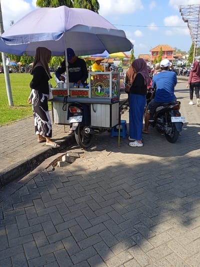 The image shows a street scene in Kebondalem, Indonesia. A food vendor operates a "Bakso" cart under a large light purple umbrella. Two customers stand on the left, while another customer is seated on a motorcycle on the right. The vendor is serving the customer while standing behind the cart. The street is paved with dark grey bricks. Behind the scene, there is a grassy area, buildings, and other motorcycles parked. The sky is blue with some clouds.
