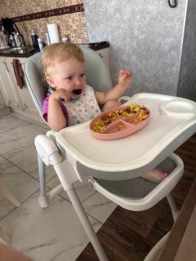A blonde toddler with blue eyes is seated in a high chair and eating from a pink divided plate. The plate contains pasta with ground meat. The child is wearing a bib and a purple shirt. The high chair is white and gray with visible legs. The background shows a kitchen area with tiled walls and cabinets. The floor is tiled with a wood-grain pattern. There is a rolled-up towel hanging on a cabinet handle to the left. A roll of paper towels is on the counter. This scene is set in the city of Kondopoga, Russia.