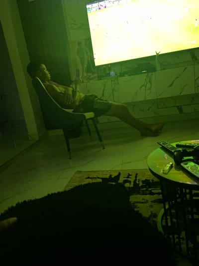 A shirtless person reclines in a black chair with their legs extended upward toward a large, bright television screen. The room is dimly lit with a green hue. A glass-topped table cluttered with remote controls sits nearby on a rug. The individual holds what appears to be a phone. The wall behind the TV has a marble pattern. A dark, obscured object fills the right side of the frame.