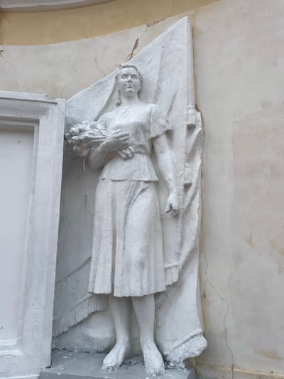 A white bas-relief sculpture depicts a woman in a knee-length dress and short-sleeved blouse, holding a bouquet of flowers in her arms. She stands against a background of a draped flag or banner. The sculpture is mounted on a textured, light-colored wall with visible cracks. To the left of the sculpture is a rectangular frame-like structure. The overall scene appears to be an exterior architectural element in Zavolzhye, Russia.