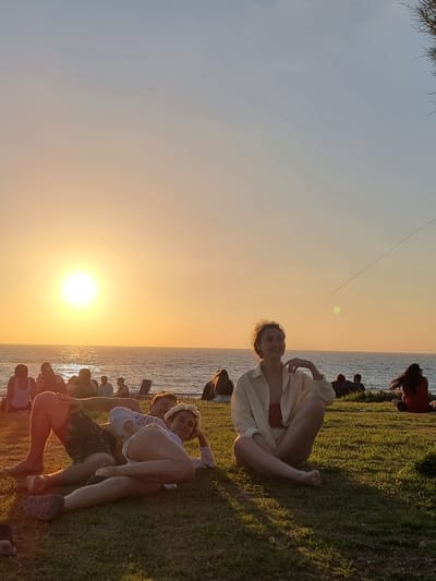The image depicts a group of people gathered on a grassy hill overlooking the Mediterranean Sea at sunset. The sun, a bright orb, is low on the horizon, casting a warm orange glow across the sky and water. In the foreground, a couple lies on the grass, embracing. To the right, a woman sits cross-legged, looking towards the sky. In the background, silhouetted figures are scattered across the hillside and along the shoreline, some sitting, others standing. The scene suggests a relaxed, communal atmosphere. The location is identified as Palmachim Beach, Israel, a popular spot known for its natural beauty and sunset views.