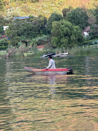 This image depicts a scene on Lake Atitlán, adjacent to the city of San Pablo La Laguna, Guatemala. In the foreground, a man is situated in a small, wooden, red-painted boat with an outboard motor mounted at the stern. He is wearing a light-colored long-sleeved shirt or jacket and a dark hat, positioned facing left (his right) with his hands resting on the boat's gunwale. The water surface exhibits ripples and reflections of green and golden light. The midground features a lush green shoreline with tall reeds along the water's edge. Two wooden dock structures are visible; one on the left appears elevated and weathered, while another on the right is partially submerged or low-lying, with a black and white speedboat moored or partially sunken nearby. Dense tropical vegetation, including banana plants, covers the land behind the docks. In the background, a steep hillside rises, densely covered in varied green foliage, patches of white flowering plants, and numerous trees. Structures are integrated into the hillside, including a building with visible solar panels on its roof towards the left and a white-walled house with a dark roof partially discernible through the trees on the right.