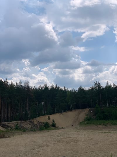 This is a landscape photograph taken in Hostomel, Ukraine. The image depicts a sandy terrain in the foreground, with a sloped sand dune rising to the left. A dense forest of pine trees forms a dark line across the horizon, separating the sand from a bright blue sky filled with dramatic, cumulus clouds. The overall scene is natural and undeveloped, suggesting a rural or semi-rural environment within the city. There are no people or specific actions visible in the image. The lighting suggests it is daytime, possibly late morning or early afternoon, with the sun illuminating the clouds from above.
