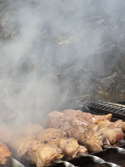 In Pidhorodne, Ukraine, skewers of marinated meat are being grilled over a fire, producing a significant amount of smoke that obscures the background. The meat, cut into uniform pieces, is skewered on metal rods and arranged on a grill. The grill itself is placed in front of a rough, stone-like surface, possibly a wall or an outdoor oven. A second grill grate is visible in the upper right portion of the image. The focus is on the meat and the smoke, suggesting an outdoor cooking event.