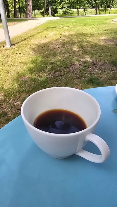 This media file features a close-up, overhead view of a white ceramic mug filled with dark coffee. The mug has a prominent handle and rests on a bright blue surface, possibly a table or chair. The background reveals a park-like setting with green grass, trees, and a paved walkway. The lighting suggests it is daytime, likely outdoors, with natural sunlight casting reflections on the coffee's surface. The scene evokes a sense of relaxation and enjoyment of a beverage in an outdoor environment. There are no people visible in the image, nor is there any visible text. The location context of Konstancin-Jeziorna, Poland, is provided.