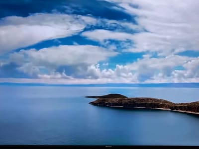 The image depicts a serene aerial view of islands in a vast body of water under a dramatic sky. The water is a deep, tranquil blue, with subtle ripples visible. Two distinct islands, characterized by their brown, textured terrain, emerge from the water. The larger island forms a crescent shape, while a smaller, conical island is situated to its left. The horizon line is a thin, hazy band separating the water from a distant landmass. Above, the sky is a vibrant spectacle of blue interspersed with voluminous white clouds, some of which are tinged with gray, suggesting depth and movement. The scene is devoid of any human presence. The branding "Hisense" is visible at the bottom edge of the image, indicating it is a photograph of a television screen. There is no information available to suggest this scene is located in the city of Ləcət, Azerbaijan.