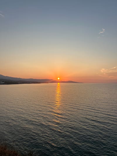 The image depicts a tranquil seascape at sunset, with the sun hovering just above the horizon. The sun casts a brilliant golden reflection across the rippling surface of the sea, creating a shimmering path of light that stretches towards the viewer. In the distance, silhouetted mountains form a rugged coastline, with a small cluster of buildings visible along the shore. The sky transitions from a soft, pale orange near the sun to a clear, pale blue at the top of the frame, with a few wispy clouds adding subtle texture.

The overall mood is serene and peaceful, evoking the quiet beauty of a day's end. The gentle undulation of the water suggests calm conditions, and the warm hues of the sunset indicate fair weather. The presence of the mountains and the coastline hints at a Mediterranean location. The image is characterized by its natural beauty and the play of light on water. No people or specific objects are discernible, focusing the attention on the grand scale of the natural landscape. The time of day is clearly sunset, and the location is likely coastal Greece, given the provided context. There is no visible text in the image.