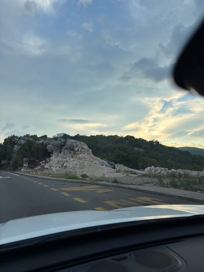 The image shows a scenic view from inside a vehicle looking out towards a rocky hillside and a cloudy sky. The scene is oriented vertically, suggesting the photo was taken from a tilted perspective.
The foreground is dominated by the interior of the vehicle, with a dark, blurred area at the top that appears to be part of the car's interior, possibly a dashboard or roof lining. To the right, a portion of the car's white bodywork and a black interior element, possibly a speaker grille, are visible. The road is visible in the lower right and extends into the middle ground. It is an asphalt road with yellow markings.
The main subjects are the natural landscape. On the left, a dramatic sky with scattered clouds is visible, with patches of bright sunlight breaking through, indicating a time of day around sunrise or sunset. The clouds are a mix of light grey and white, with some darker, more ominous clouds present.
The middle ground features a steep, rocky hillside covered in dense green vegetation. Large, light-colored rocks are prominent, with trees and shrubs growing amongst them. A small, white building is partially visible nestled on the hillside amongst the rocks and trees. The hillside appears somewhat rugged and natural, with evidence of erosion or rockfalls.
The background consists of a further stretch of forested hillside, rising towards a hazy horizon. The overall impression is of a natural, somewhat wild landscape.
There is no clear activity or event taking place, the image appears to capture a moment in time during a journey. The weather seems mixed, with sunshine and clouds present. The location context suggests Brežine, Montenegro, which aligns with the rocky, mountainous terrain and vegetation depicted. No legible text is visible in the image.