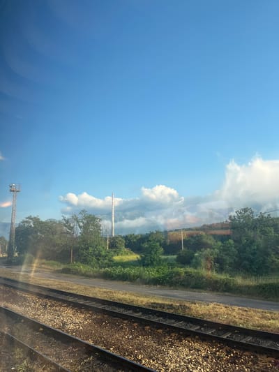 The image displays a pastoral landscape viewed from a moving train. In the foreground, two sets of train tracks are visible, filled with ballast. Beyond the tracks, a dirt road runs parallel to the railway. The middle ground features a lush green field bordered by a dense line of trees and bushes. In the background, a tall, striped industrial chimney and a metallic observation tower stand against a bright blue sky dotted with white, fluffy clouds. Hills are faintly visible in the far distance, shrouded in mist. The scene is bathed in natural sunlight. The location is Makotsevo, Bulgaria.