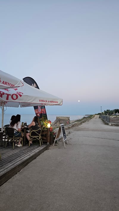 The image depicts an outdoor seating area of a restaurant or cafe near the Black Sea coast in Shabla, Bulgaria. The scene is set at dusk, with a pale blue sky and a full moon visible. In the foreground, a wooden deck with tables and chairs is occupied by three individuals. They are seated and appear to be engaged in conversation or dining. One person is wearing a white t-shirt and denim shorts, another is wearing a black t-shirt, and the third's attire is partially obscured. A large white umbrella with "STELLA ARTOIS" branding covers part of the seating area. A decorative light fixture illuminates some flowers. A chalkboard sign stands near the deck, its text illegible. To the right of the seating area, a concrete path leads away from the viewer, curving towards the coast. The path is bordered by a low wall and vegetation. In the distance, the sea stretches to the horizon, with a concrete structure, possibly a remnant of a pier or building, visible on the coastline. The overall atmosphere suggests a tranquil evening by the sea.