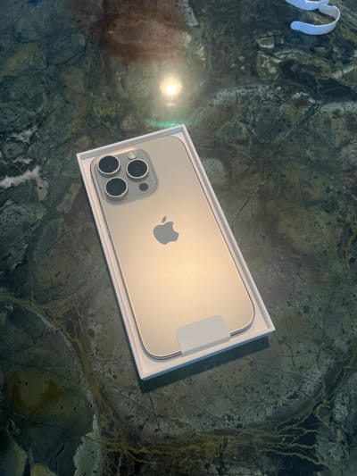 An unboxed, light gold or silver iPhone rests in its white packaging tray on a polished, dark green or black stone surface with light veining. The iPhone, oriented vertically, displays its rear panel, featuring the centered Apple logo and a prominent triple-camera array with three large lenses, a smaller sensor, and a flash module in the upper left quadrant. A white pull tab is visible at the bottom of the phone within the tray. A bright, circular light reflection is present on the stone surface above the phone, indicating an overhead light source. A white, looped fabric strap is partially visible in the upper right corner of the frame. The scene is located within the city of Hornsea, United Kingdom. No individuals are present.